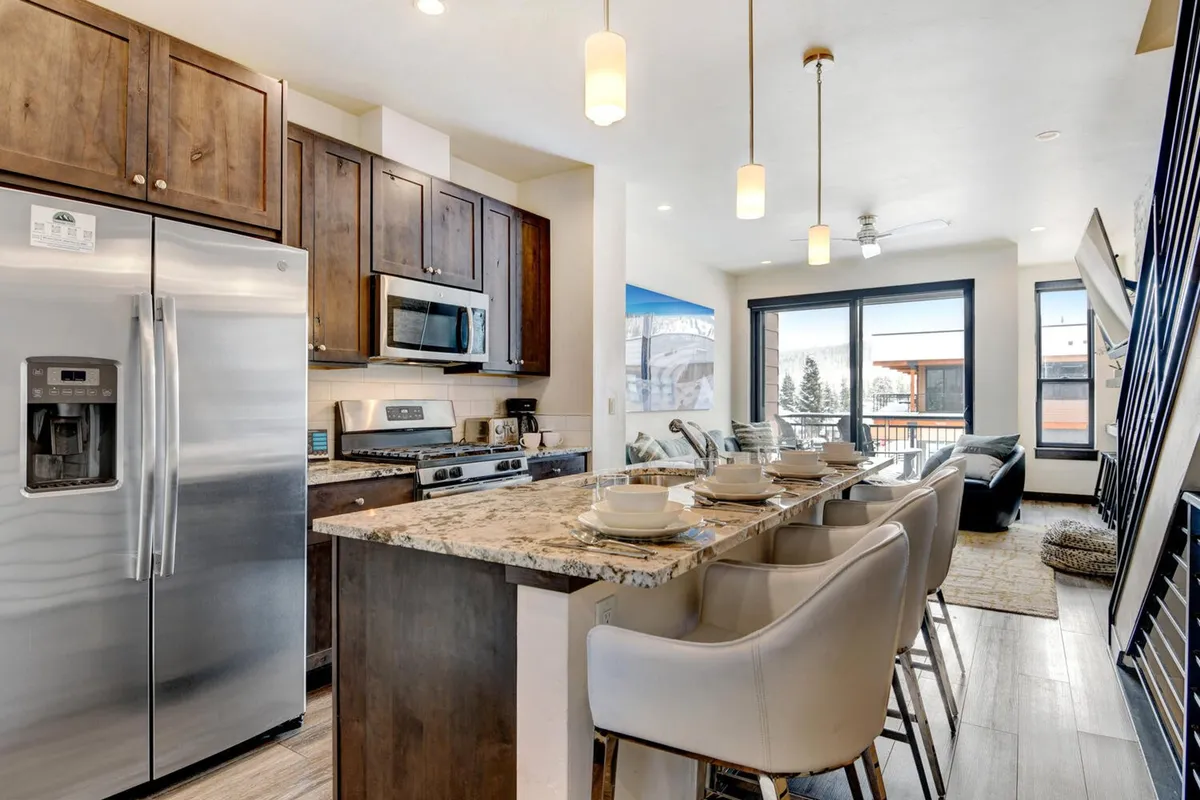 A bright, modern kitchen with a granite island that flows seamlessly into a dining space with ample seating. Stainless steel appliances and large windows contribute to an inviting atmosphere, ideal for socializing and shared meals.