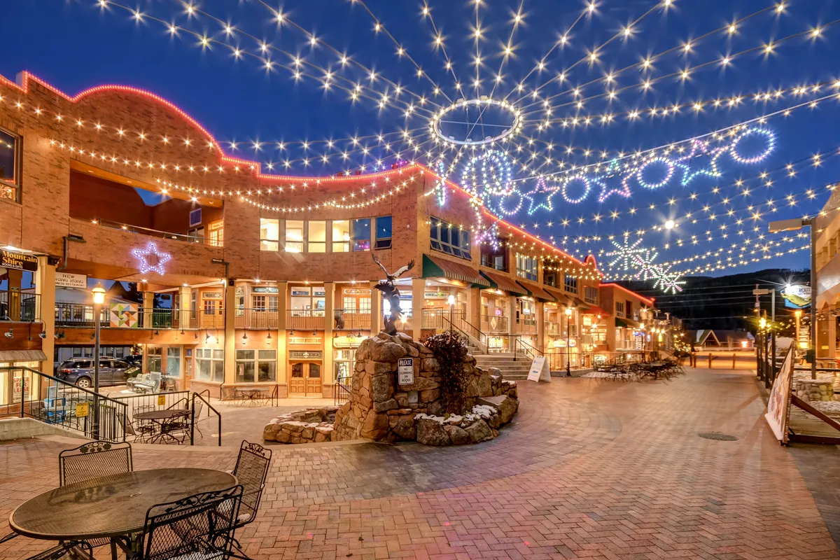 A charming village square illuminated by twinkling lights strung overhead, with decorated buildings and a central fountain. People can be seen strolling, dining, shopping, and socializing, capturing the vibrant, festive atmosphere of Winter Park’s local attractions.