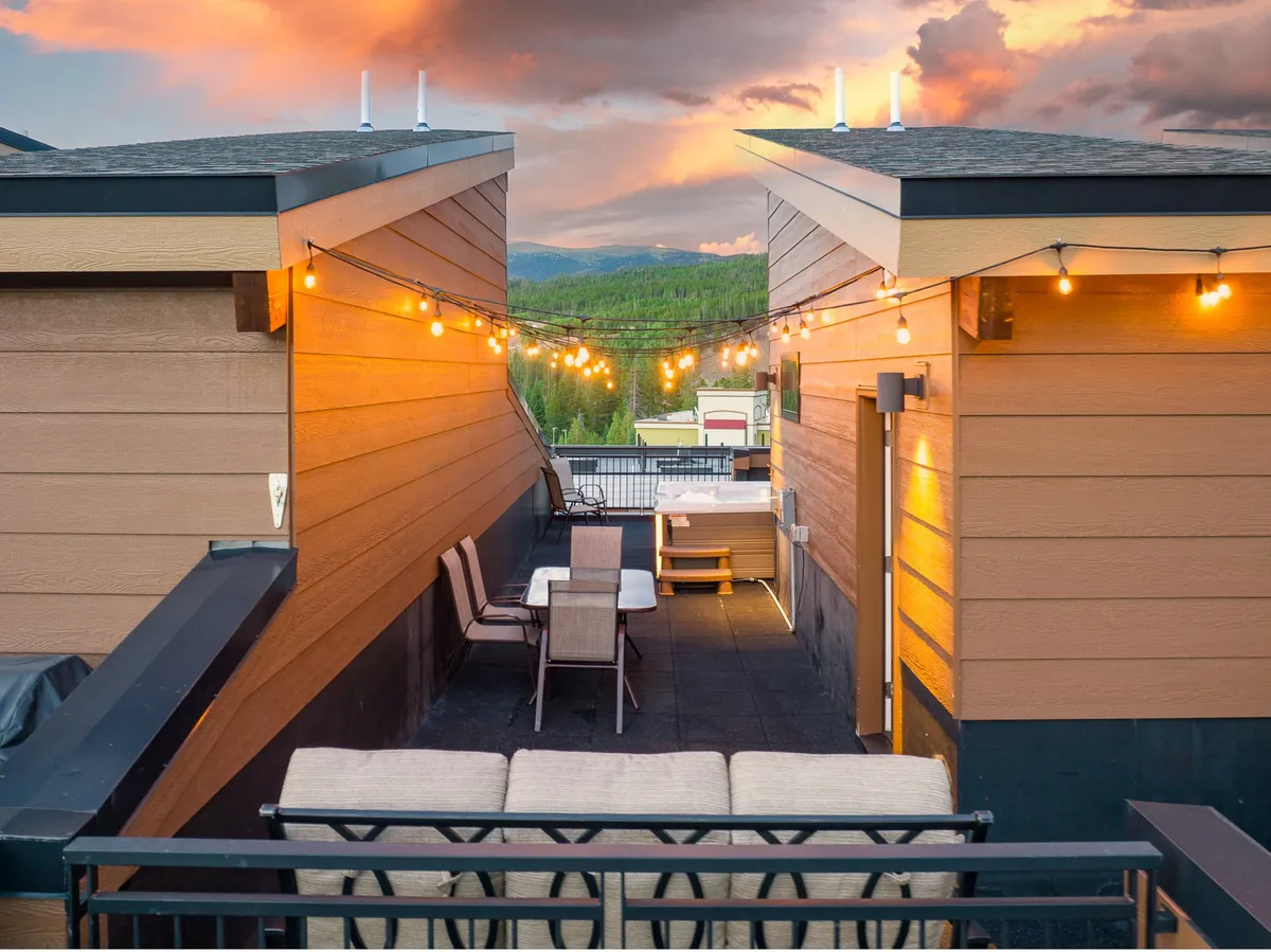 An outdoor deck framed by string lights and comfortable seating, overlooking a stunning mountain view. The setup includes lounge areas and a patio table, inviting both relaxation and outdoor dining while soaking in nature's beauty.