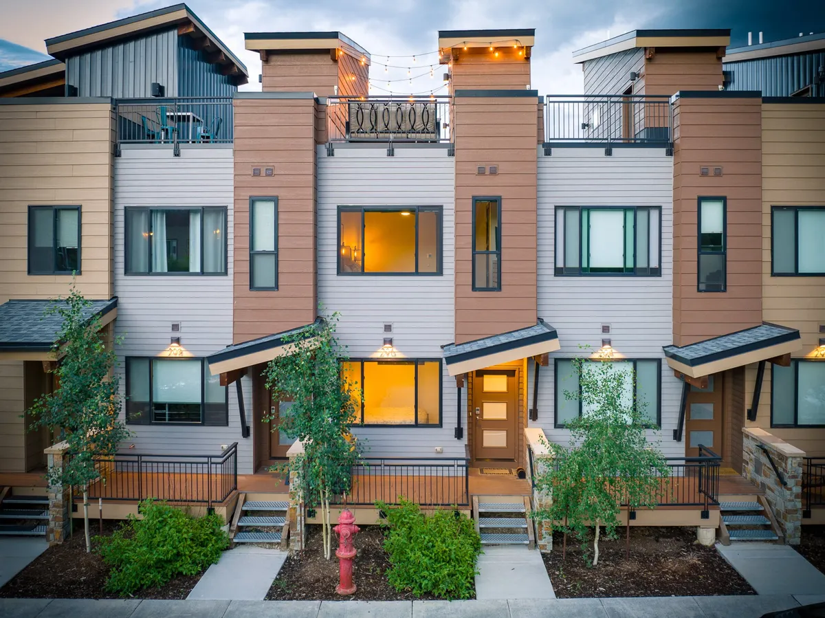The image showcases the villa's striking modern architecture, with an inviting outdoor patio and lush landscaping. The balcony is adorned with glowing string lights, offering a warm ambiance perfect for evening gatherings, and the sunset in the background enhances the welcoming, lively mood.