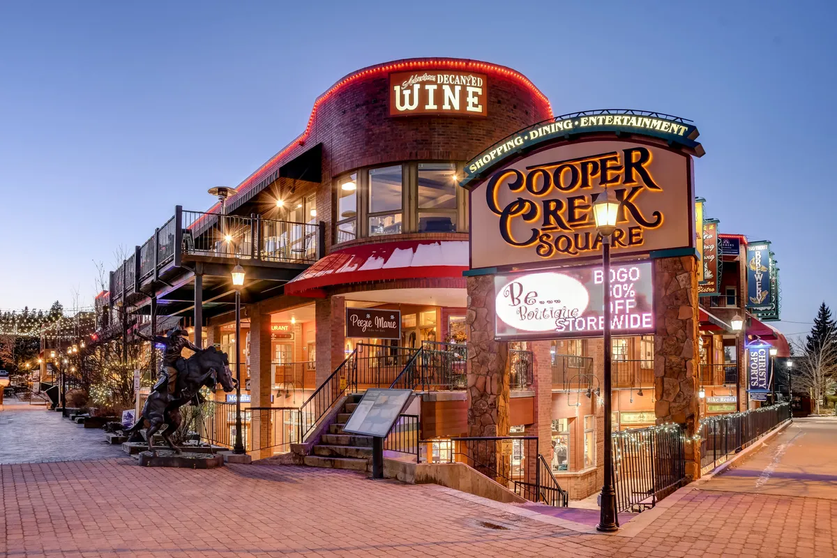 The image displays the vibrant entrance to Cooper Creek Square, featuring festive outdoor seating, decorative lighting, and visible storefront signs for wine shops and eateries. The atmosphere is lively, suggesting a hub of activity, dining, and entertainment in downtown Winter Park.