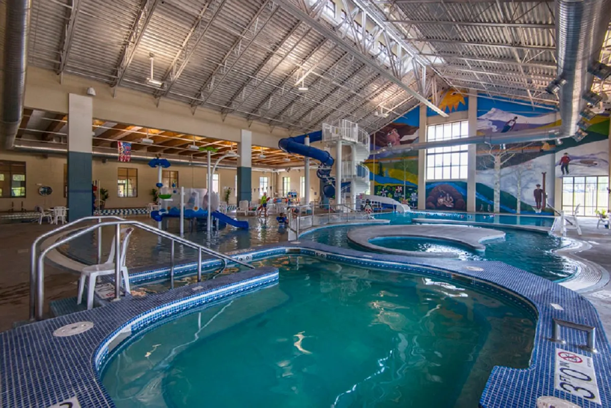 A joyful, colorful indoor pool scene at the Grand Park Rec Center, with children enjoying the water slide, families gathering by the pool, and a vibrant mural setting a playful mood.