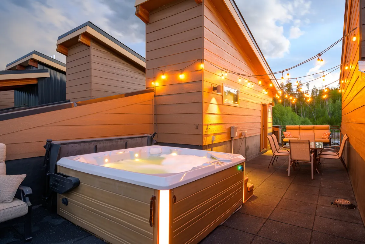 A private rooftop patio featuring a bubbling hot tub, comfortable outdoor seating, and string lights overhead. The scene is set for evening relaxation, with the inviting glow of lights hinting at a cozy and festive atmosphere.