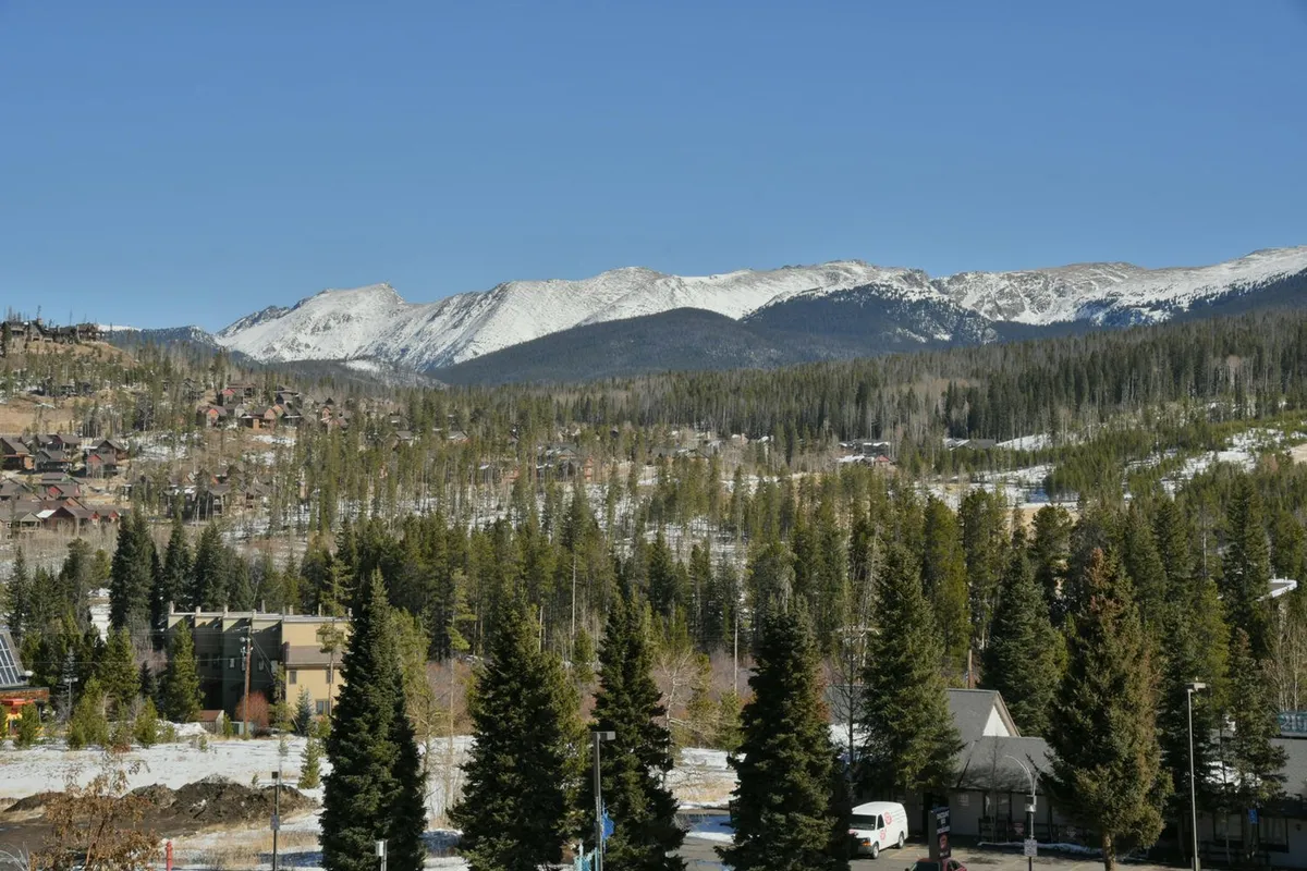 An expansive view of snow-capped mountains and dense pine forests with small buildings in the foreground. The serene landscape evokes the tranquility and beauty of the Winter Park area, ideal for outdoor activities or peaceful contemplation.