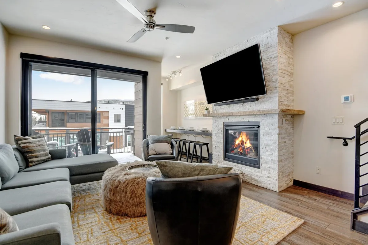 A spacious open-plan living room featuring a plush sofa facing a modern gas fireplace, with a wall-mounted HD smart TV above. Large windows let in natural light, offering glimpses of the outdoors, while accent decor creates a warm, welcoming atmosphere.