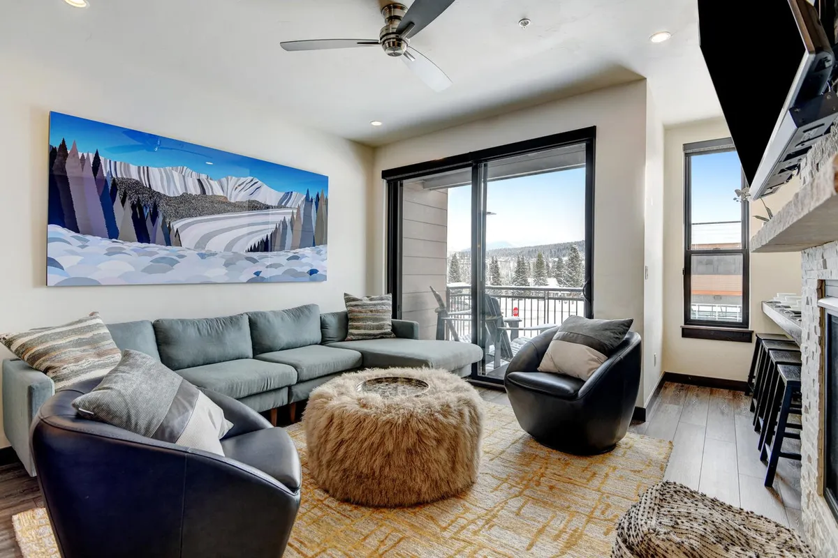 A contemporary living room with a cozy sectional sofa and a furry ottoman, accented by a striking abstract painting. Large windows provide outdoor views and fill the space with light, while the arrangement encourages socializing and group activities like board games.