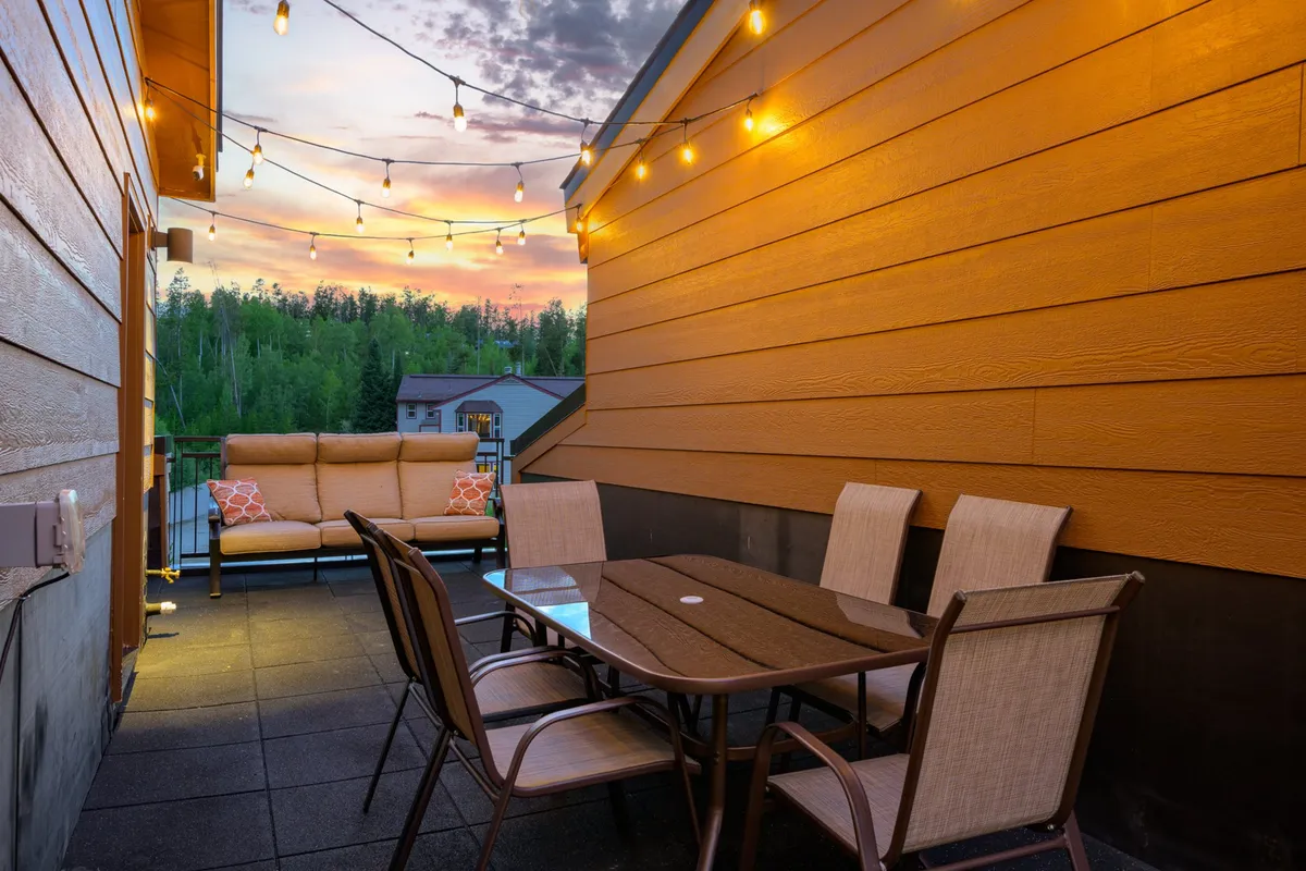 A welcoming outdoor dining area with string lights, cozy seating, and a table set for an evening gathering. Surrounded by trees and suffused with warm light, the space invites socializing and leisurely meals as the sun sets.
