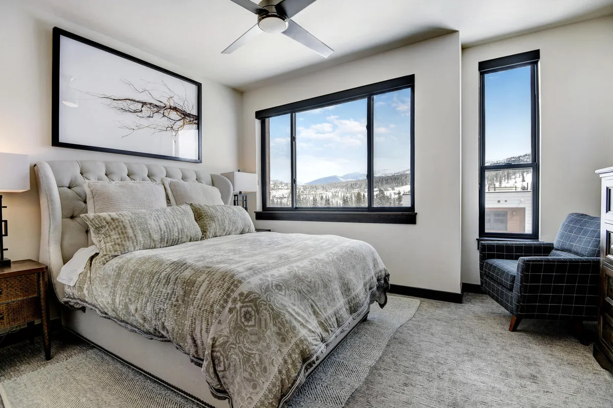A tranquil bedroom featuring a queen bed with decorative bedding, large windows offering views of the mountains, stylish wall art, and a cozy armchair. The room is bathed in soft natural light, creating a calming, restful atmosphere perfect for unwinding after a long day.